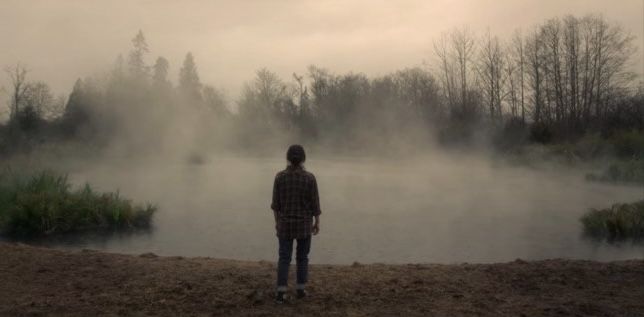 a man standing in front of a body of water on a foggy day with trees behind him