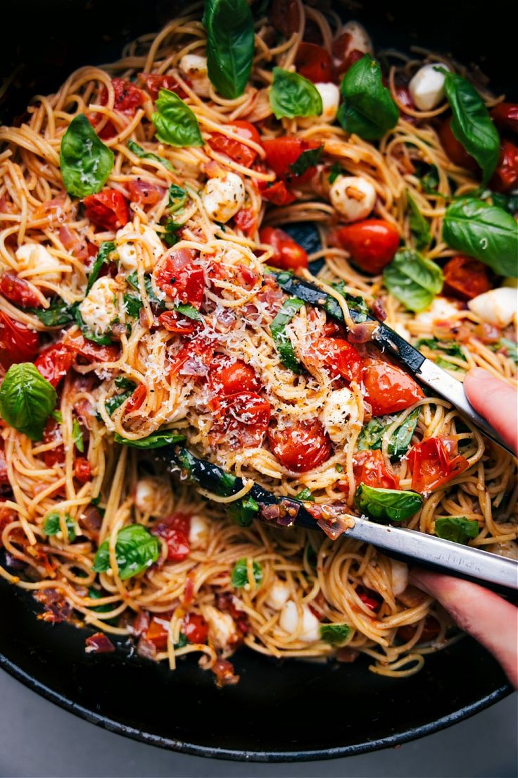 someone is holding a fork over a plate of spaghetti with tomatoes, basil and mozzarella