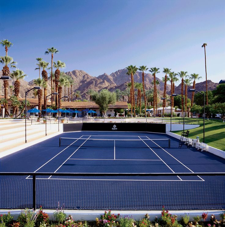 an outdoor tennis court surrounded by palm trees and mountains in the background with blue skies