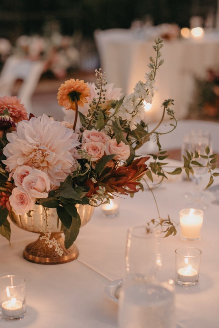 large sized vase overflowing with blooms as a wedding table centerpiece surrounded by small candles Fall Dahlia Centerpiece, Mixed Wedding Centerpieces, Ranaculus Wedding Centerpieces, Circle Table Floral Centerpiece, Copper Vase With Flowers, Dahlia Table Centerpieces, Winter Floral Centerpieces For Table, Terracotta Wedding Floral Arrangements, Dahlias Wedding Centerpieces