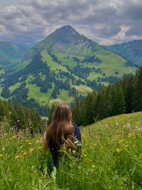 a woman sitting on top of a lush green hillside covered in flowers next to mountains