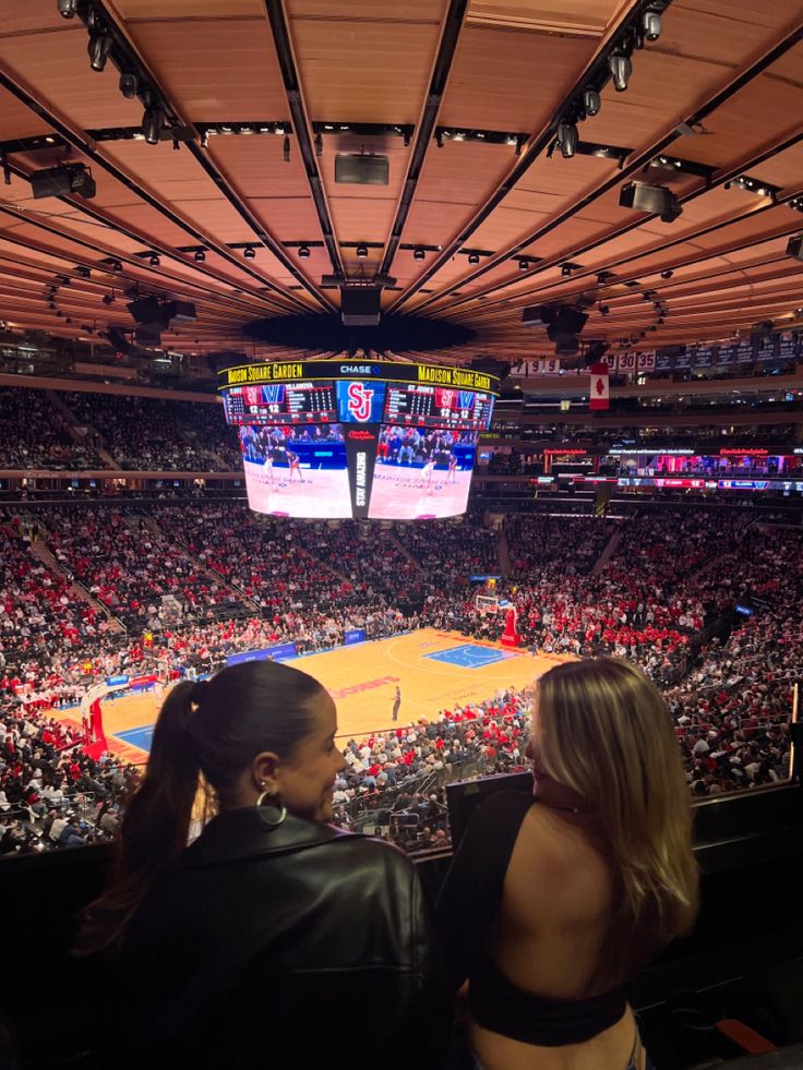 two women sitting in front of an arena watching a basketball game on the big screen