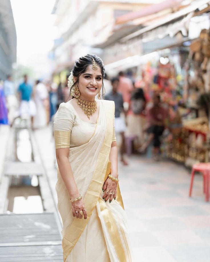 a woman in a white sari and gold jewelry standing on a street side walk