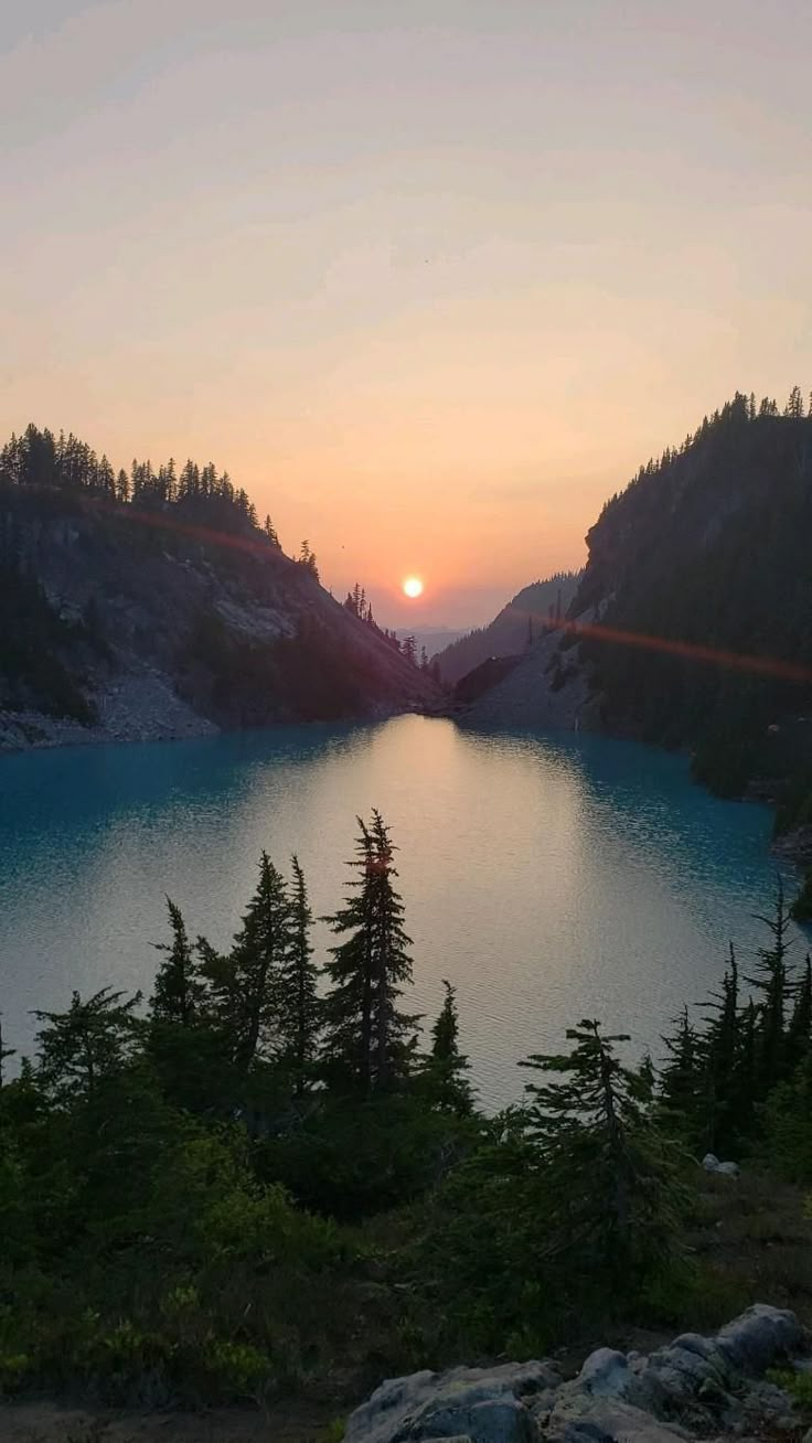 the sun is setting over a lake surrounded by mountains and pine trees in the foreground