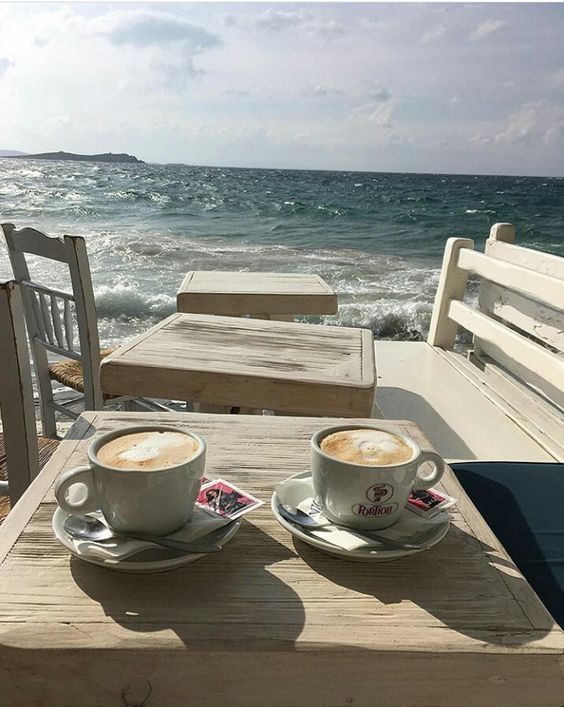 two cups of coffee sitting on top of a wooden table next to the ocean and beach