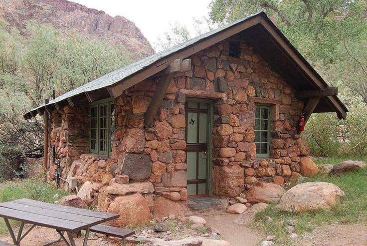 a small stone building with a picnic table in front of it and mountains behind it