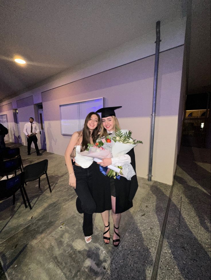 two women in graduation gowns holding flowers and posing for the camera with one another