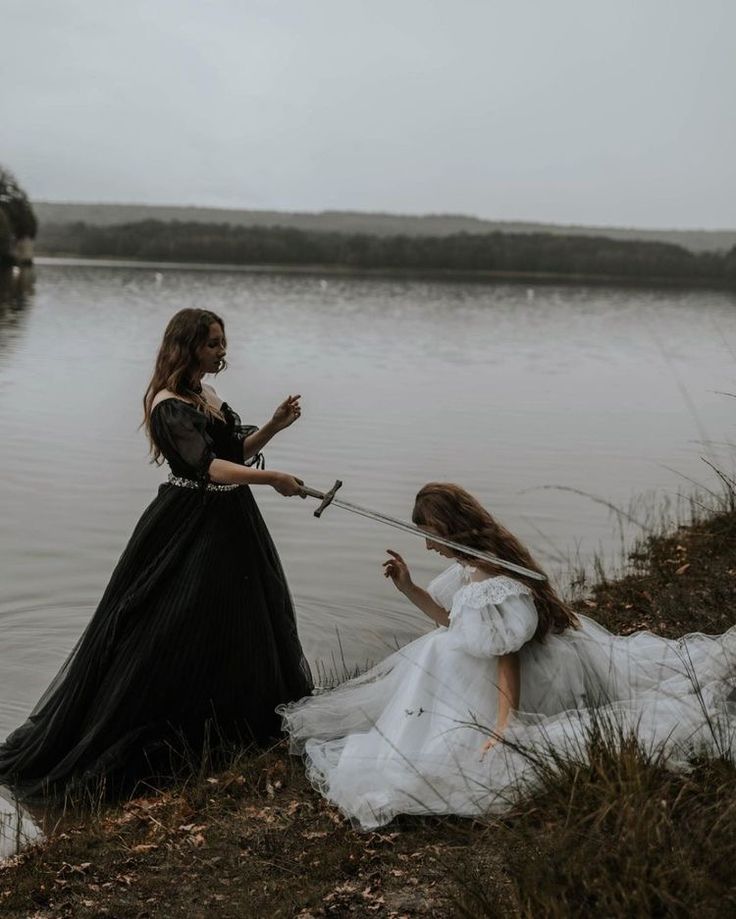 two women dressed in black and white are sitting on the bank of a lake with swords