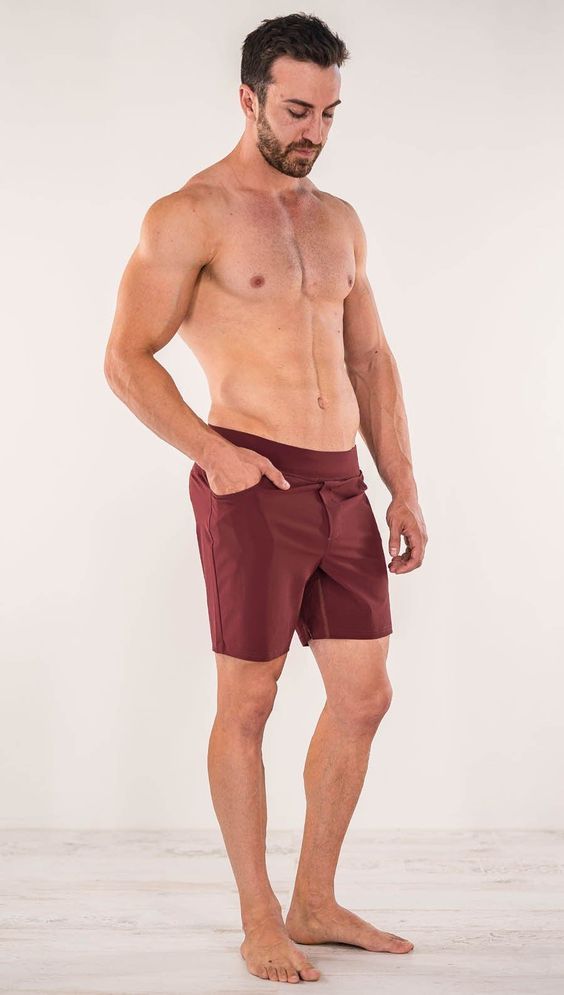 a man with no shirt is standing in front of a white wall and wearing maroon swim trunks