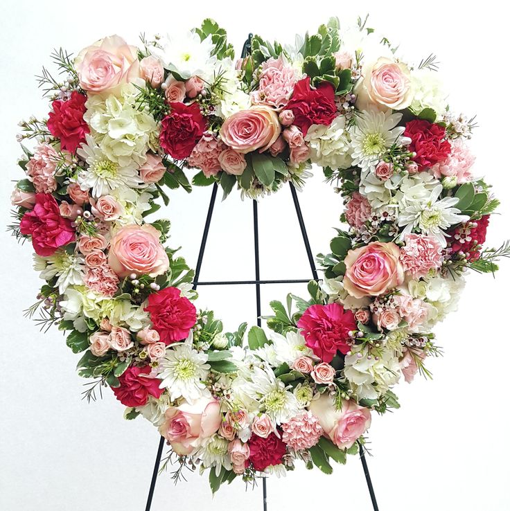 a heart shaped wreath with pink and white flowers on a black stand against a white background