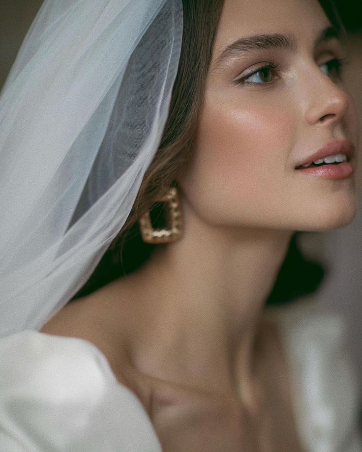 a woman in a wedding dress with a veil over her head and wearing gold earrings