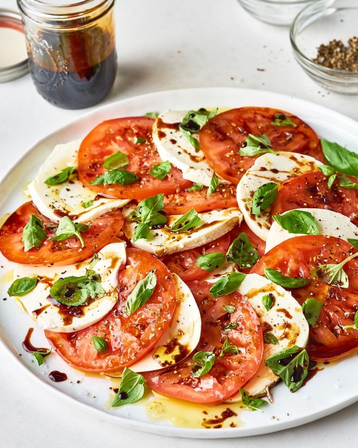 tomatoes and mozzarella on a white plate next to jars of olives, seasoning and pepper