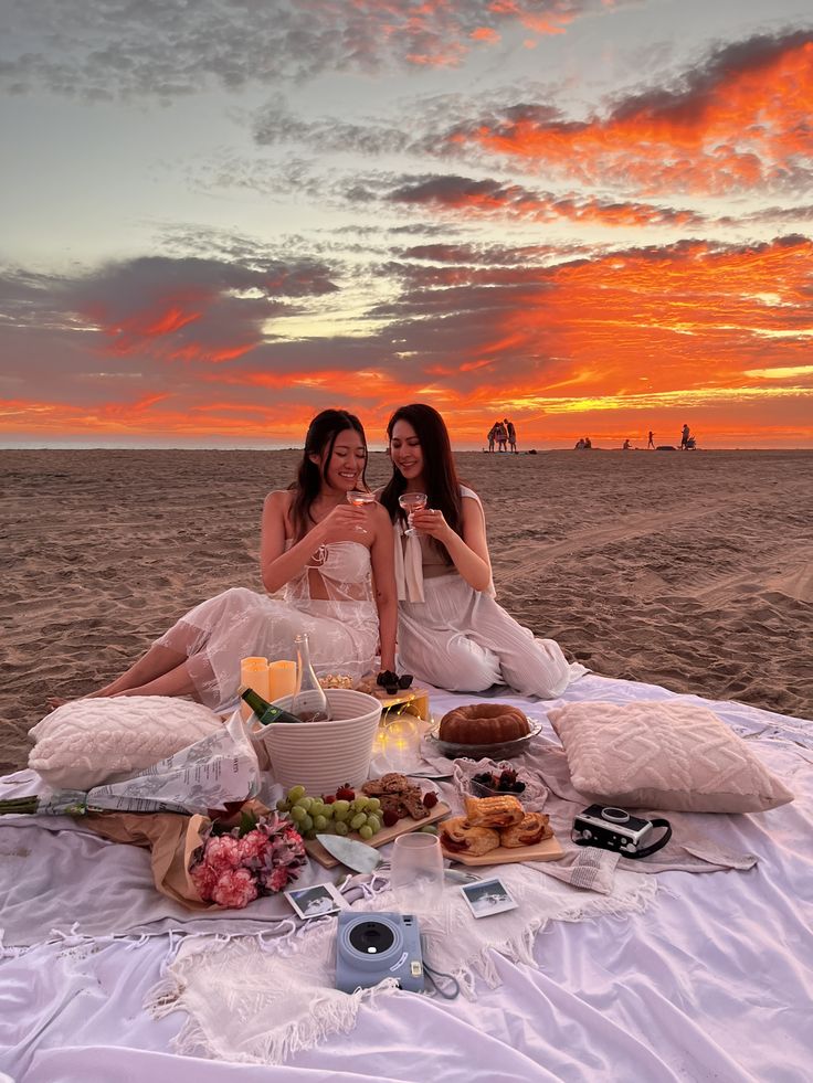 two women sitting on the beach at sunset toasting with wine glasses in their hands