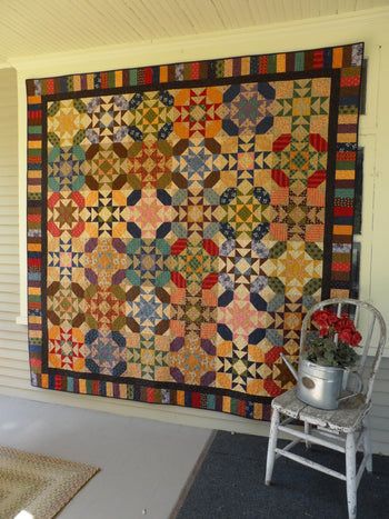 a quilt hanging on the side of a house next to a chair and flower pot