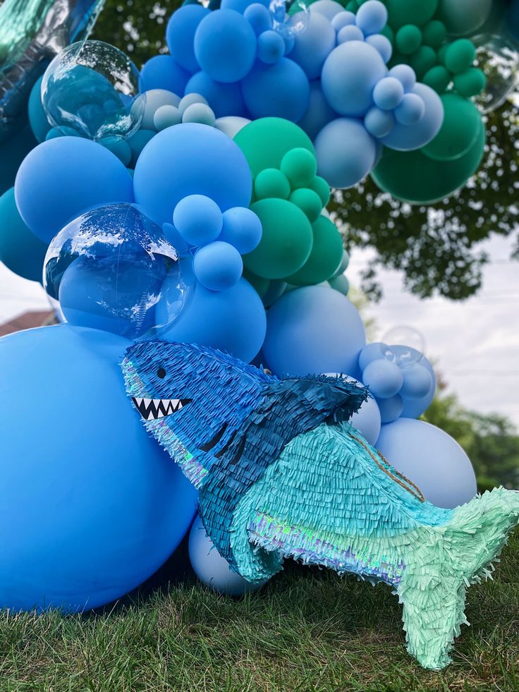 an assortment of blue and green balloons in the shape of a fish, under a tree