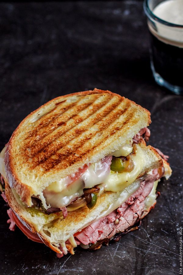 a roast beef sandwich with melted cheese and pickles on a black surface next to a glass of milk