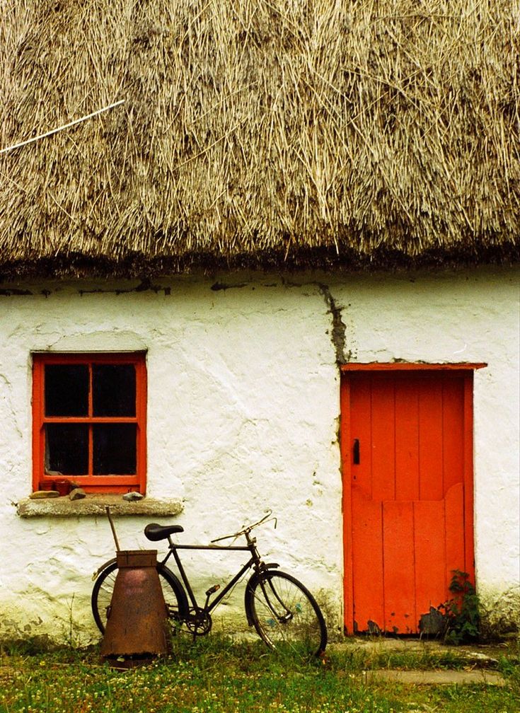 a bicycle parked in front of a thatched roof house with red door and window