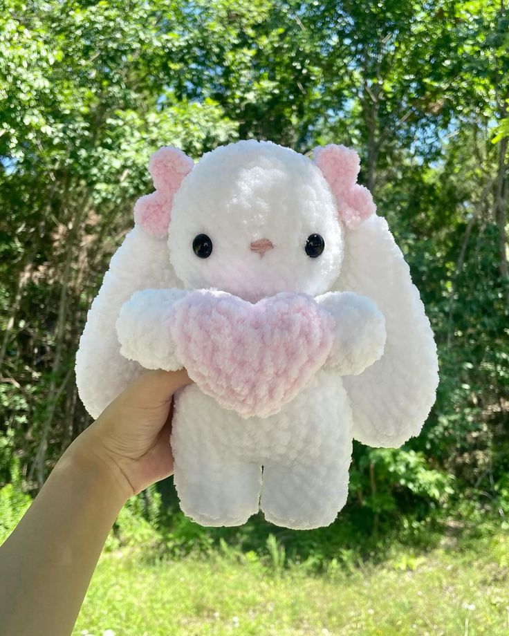 a hand holding a small white stuffed animal with pink ears and eyes, in front of some trees