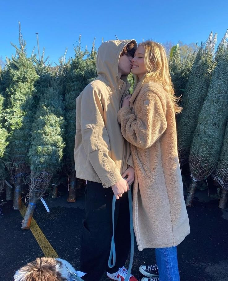 a man and woman kissing while walking their dog in front of christmas trees at a tree farm