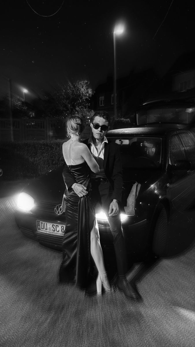 a man and woman standing next to each other in front of a car at night