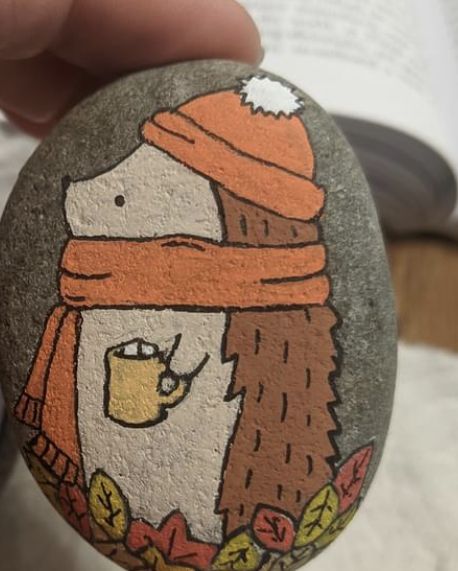 a hand holding a painted rock with an eagle wearing a hat and scarf on it