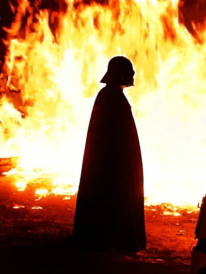 a man standing in front of a large fire with a hat on and cape over his head