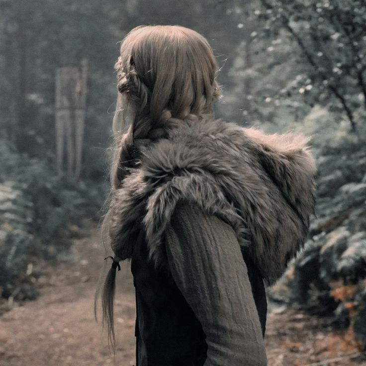 a woman with long hair walking down a path in the woods wearing a fur vest