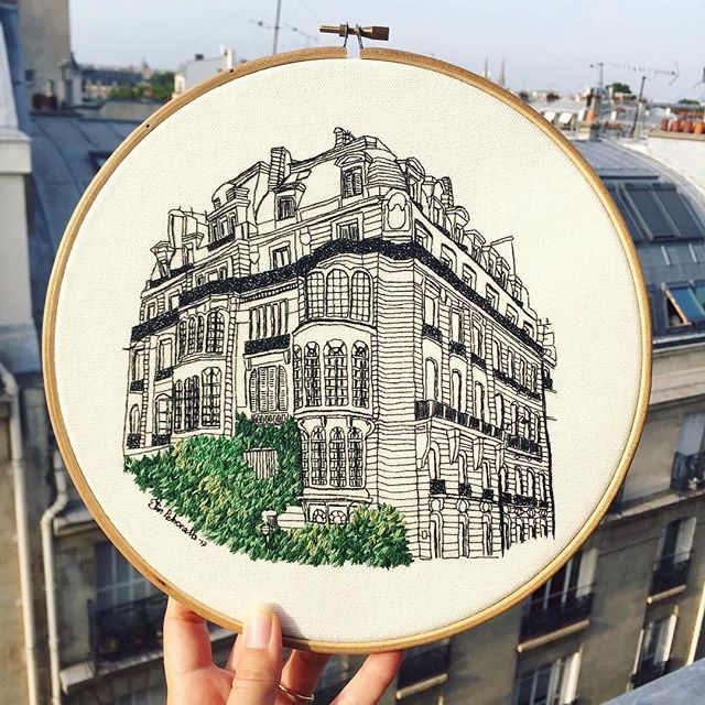 a person holding up a embroidery project with a building in the background and trees on it