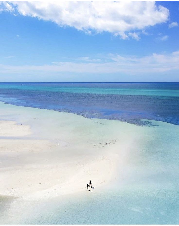 two people are walking on the beach in front of an ocean with blue sky and white sand