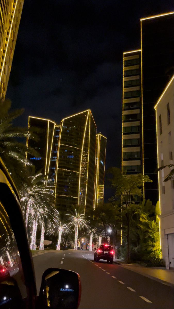 cars driving down the street at night with tall buildings in the backgrouds