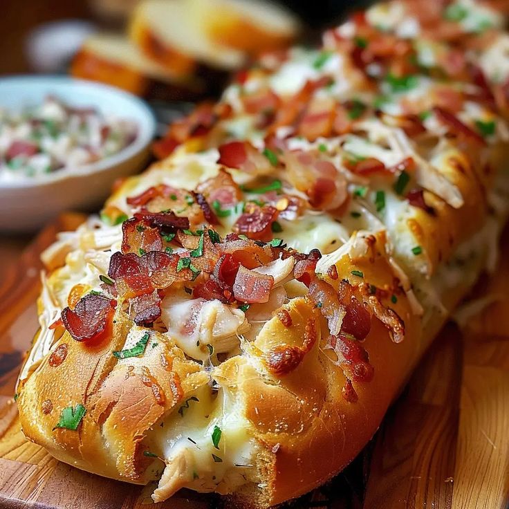 a long piece of bread covered in bacon and cheese on a cutting board next to a bowl of salad