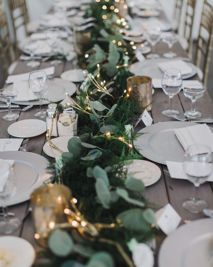 a long table is set with white plates and silverware, greenery and candles