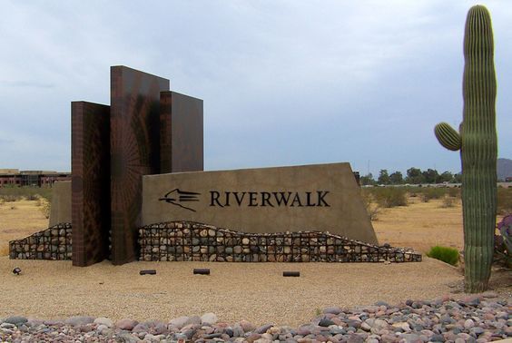 a sign that says riverwalk in front of a cactus and some rocks on the ground