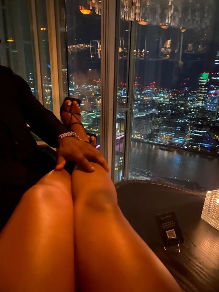 a woman's legs are shown in front of a window overlooking the city at night