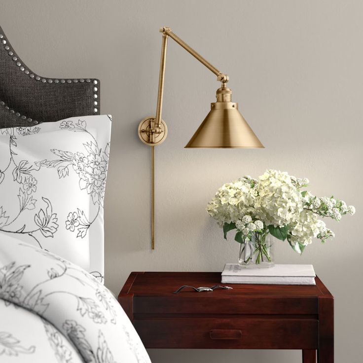 a nightstand with flowers on it next to a lamp and bed in a room that has white walls