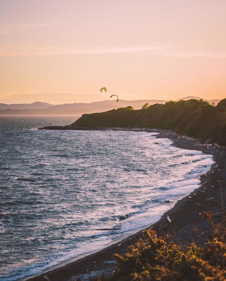 two parasailers are flying over the water at sunset on an ocean shore with mountains in the background