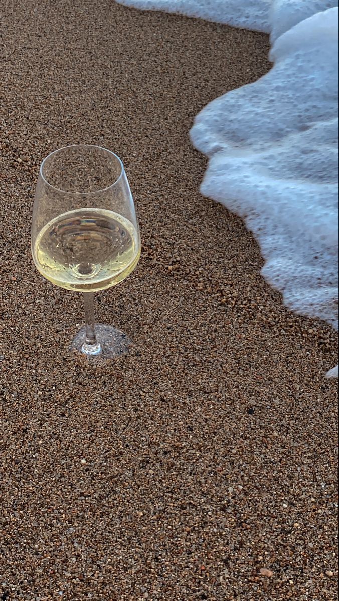 a glass of wine sitting on top of a sandy beach next to the ocean waves