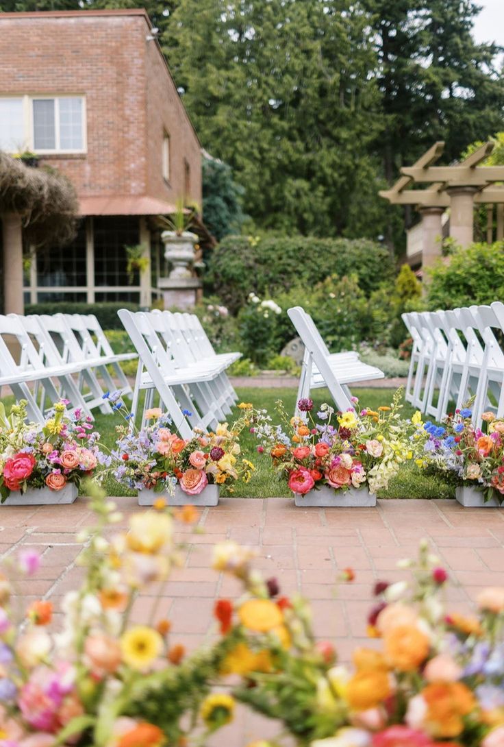 rows of white folding chairs with colorful flowers on them in front of a brick building