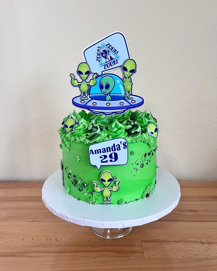 a green cake with aliens on it sitting on top of a wooden table next to a white wall