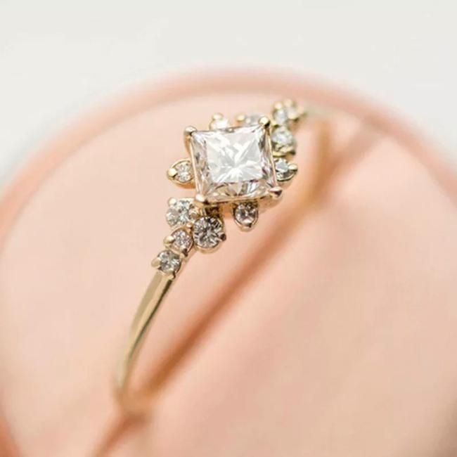 an engagement ring with a princess cut diamond surrounded by small white and clear stones on a pink box