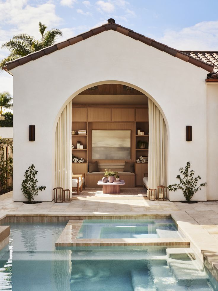 an outdoor swimming pool in front of a white stucco house with arched doorway and windows