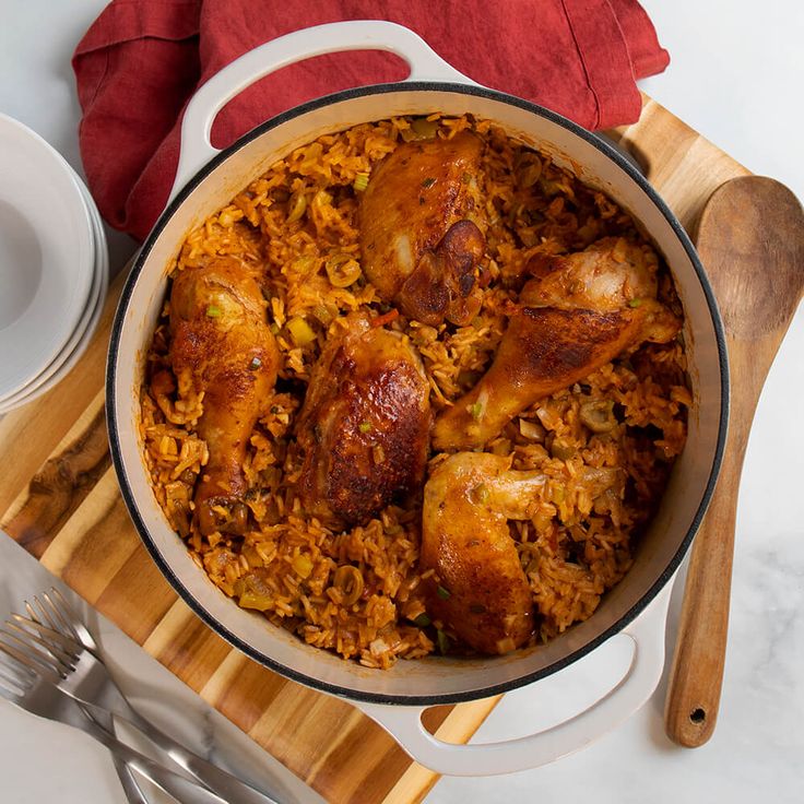 a large pot filled with rice and chicken on top of a wooden cutting board next to silverware