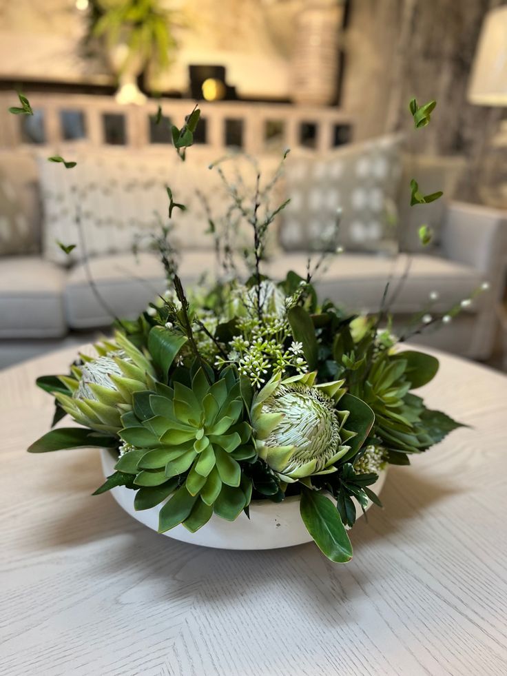 a white table topped with a vase filled with green plants and flowers next to a couch