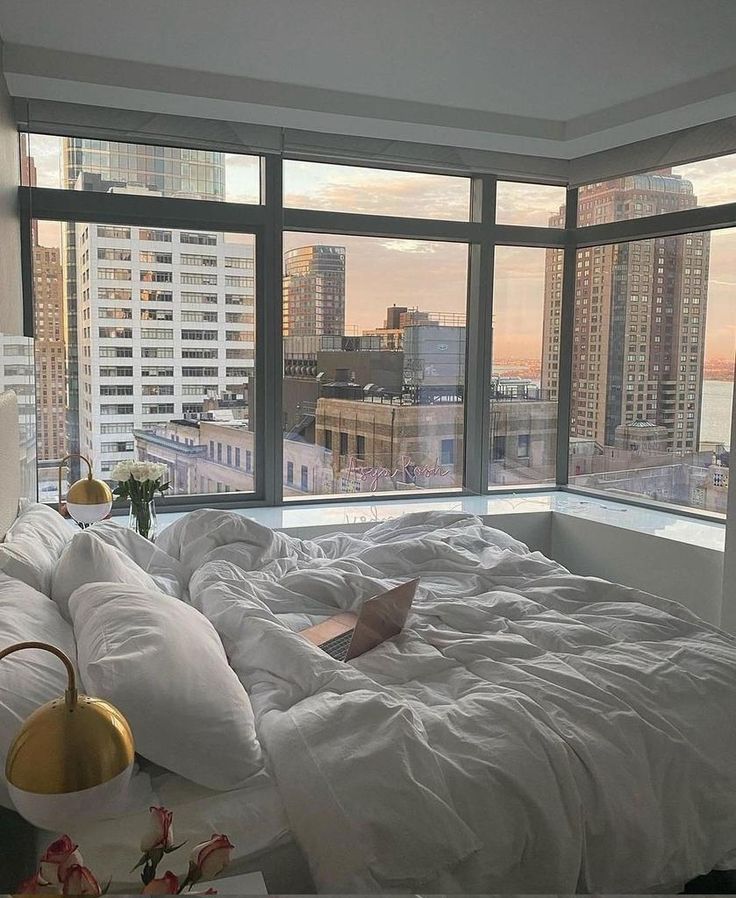 an unmade bed with white sheets and pillows in front of large windows overlooking the city