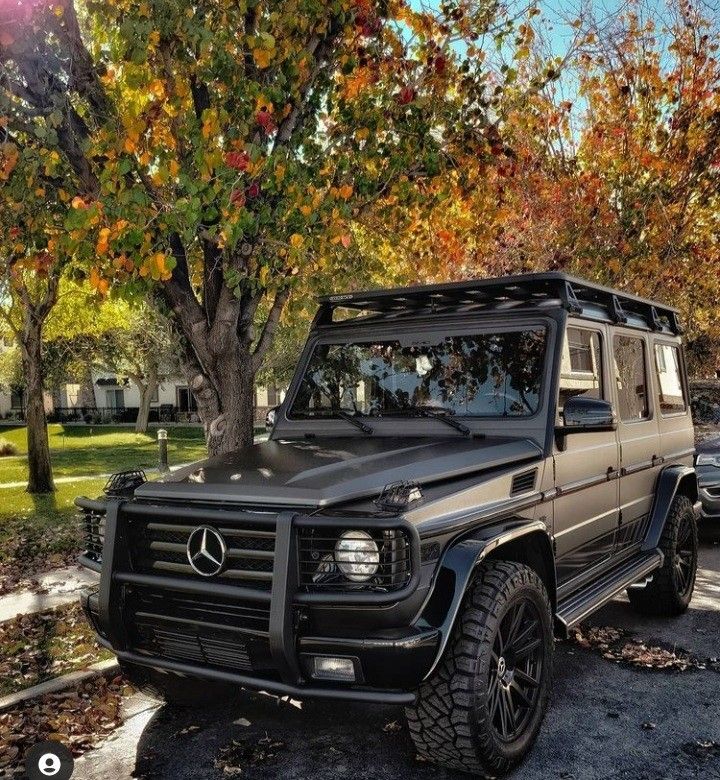 a mercedes g - class parked in front of some trees with leaves on the ground