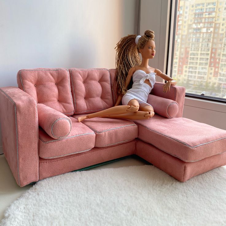 a barbie doll sitting on top of a pink couch in front of a large window