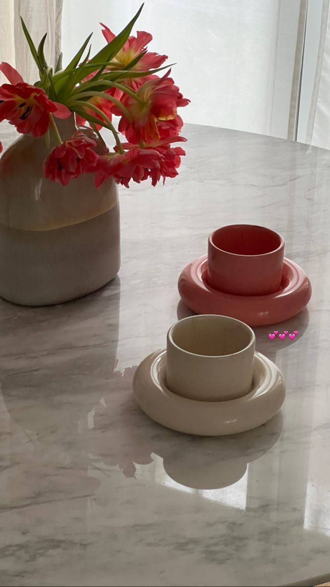 two cups and saucers sitting on a table with flowers in the vase behind them