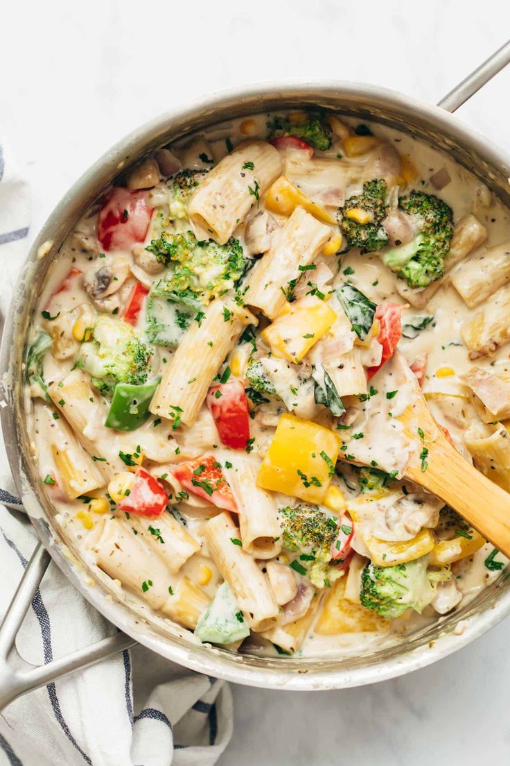 a pan filled with pasta and vegetables on top of a white table cloth next to a wooden spoon