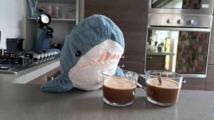 a stuffed shark sitting next to two cups of coffee on a kitchen counter with an oven in the background