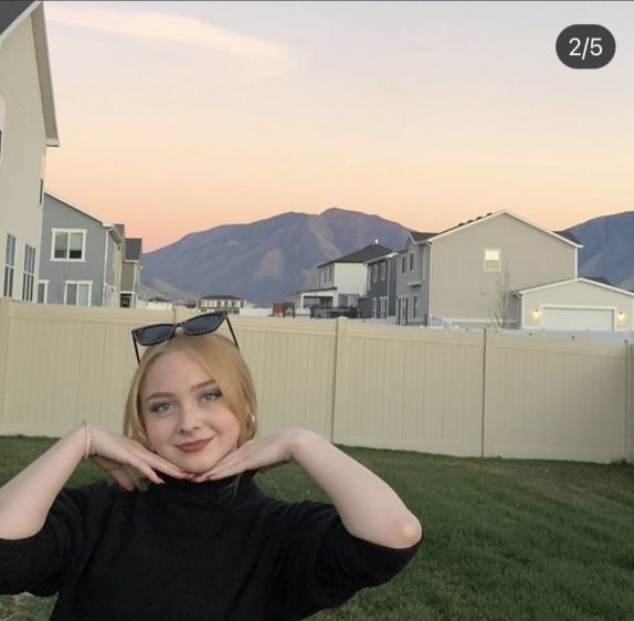 a woman is posing in front of a fence with her hands behind her head and mountains in the background